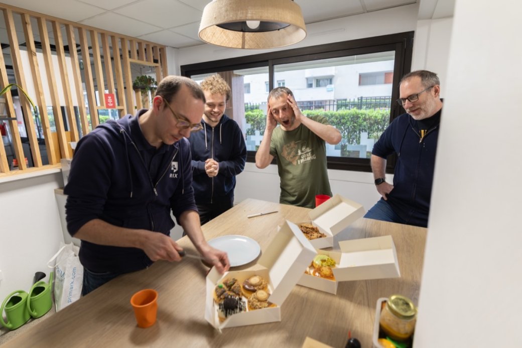 Quatre membres de l'équipe Rix réunis autour d'une table de bureau couverte de pâtisseries, l'un découpant une part, un autre l'air faussement horrifié